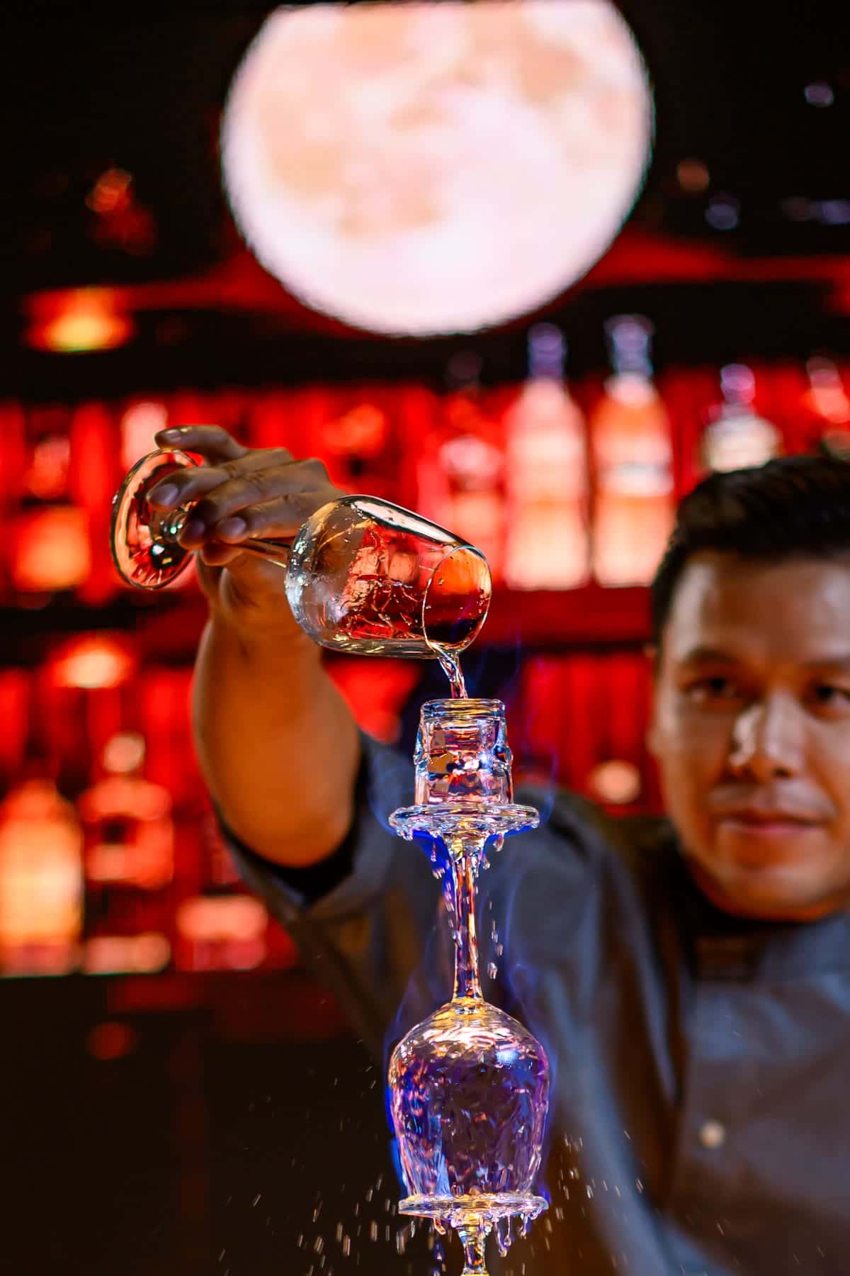 A bartender pouring a signature cocktail at the counter, the lit bottle wall behind him