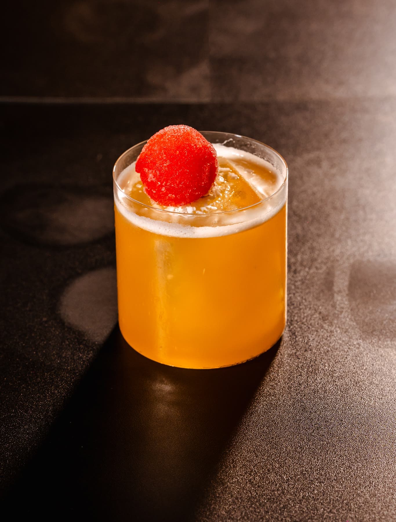 An amber cocktail in a rocks glass topped with a red sugared sphere, on a dark table