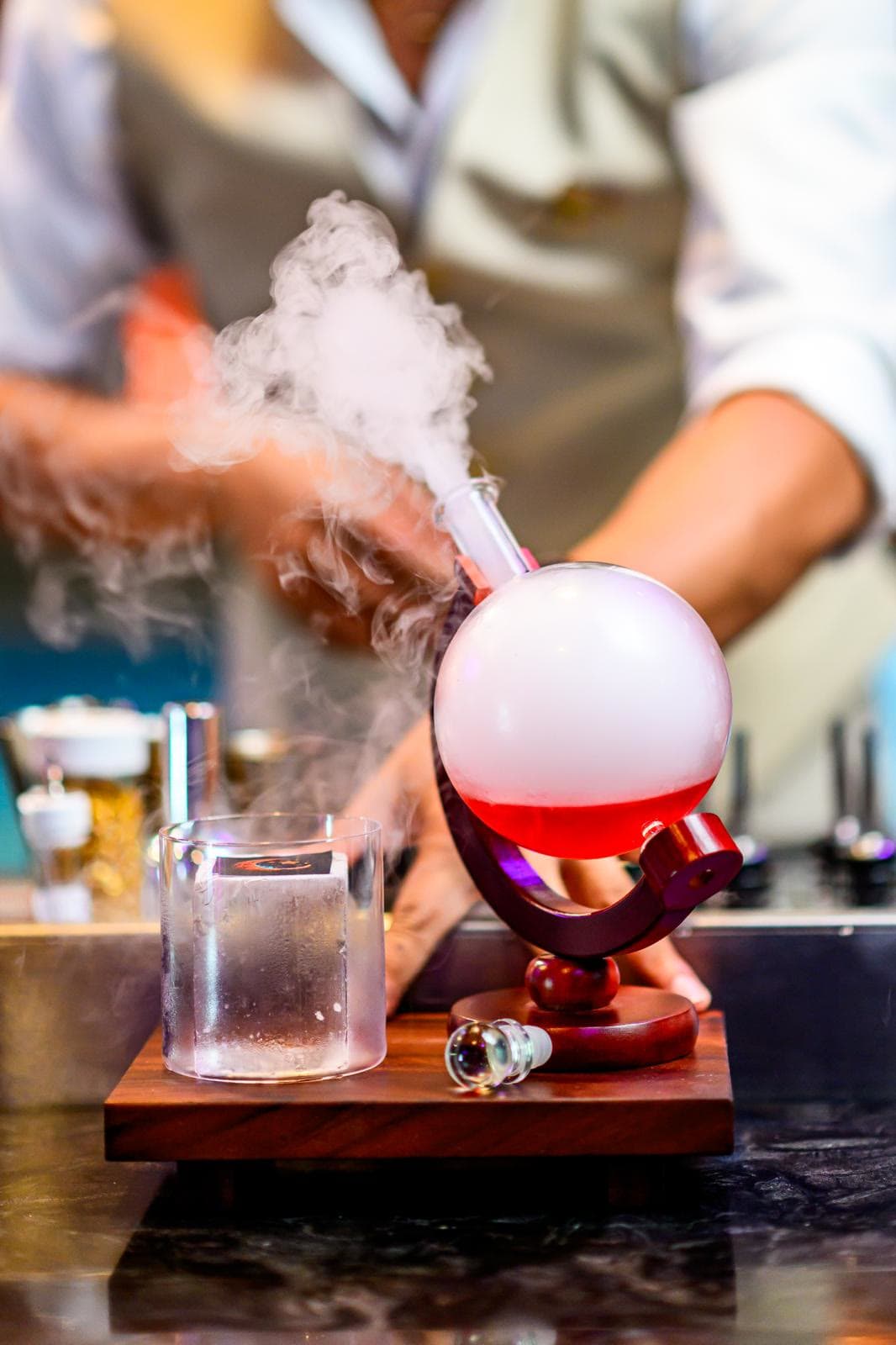 A red globe decanter on a wooden stand pouring white vapour over the bar as the bartender works behind it