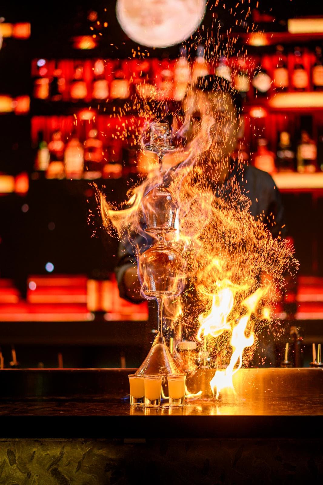 A column of flame rising from the bar top in front of the lit spirits wall, sparks in the air