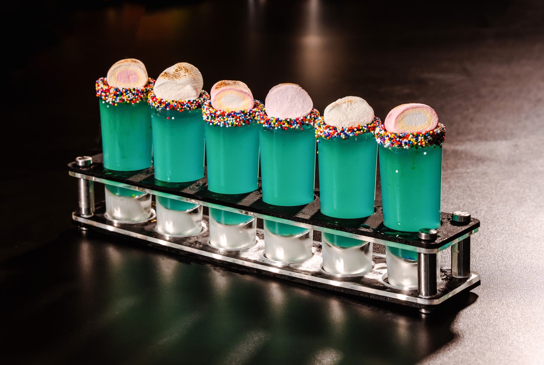 A row of blue shots rimmed with rainbow sprinkles, a toasted marshmallow on each