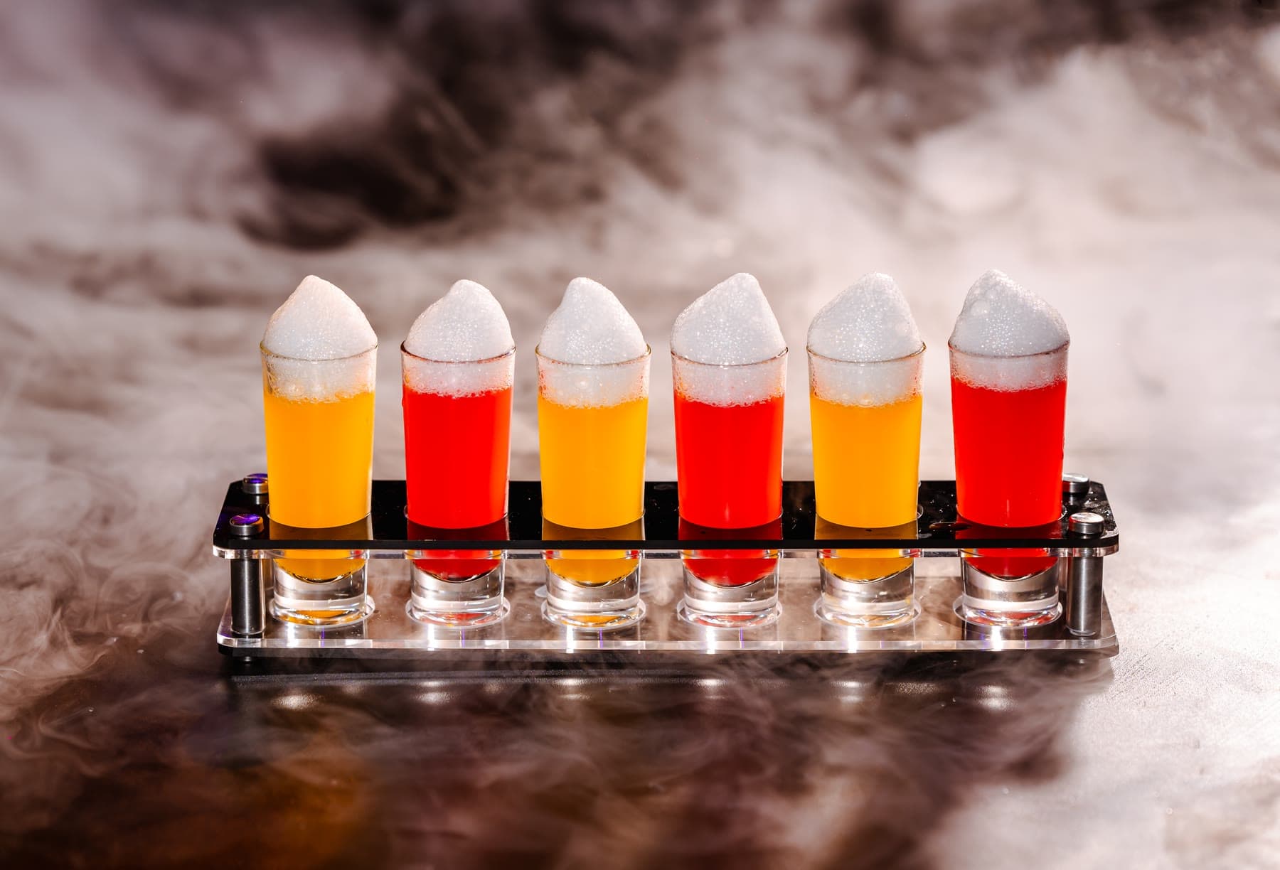 A row of six shot glasses, orange and red, each under a cap of white foam