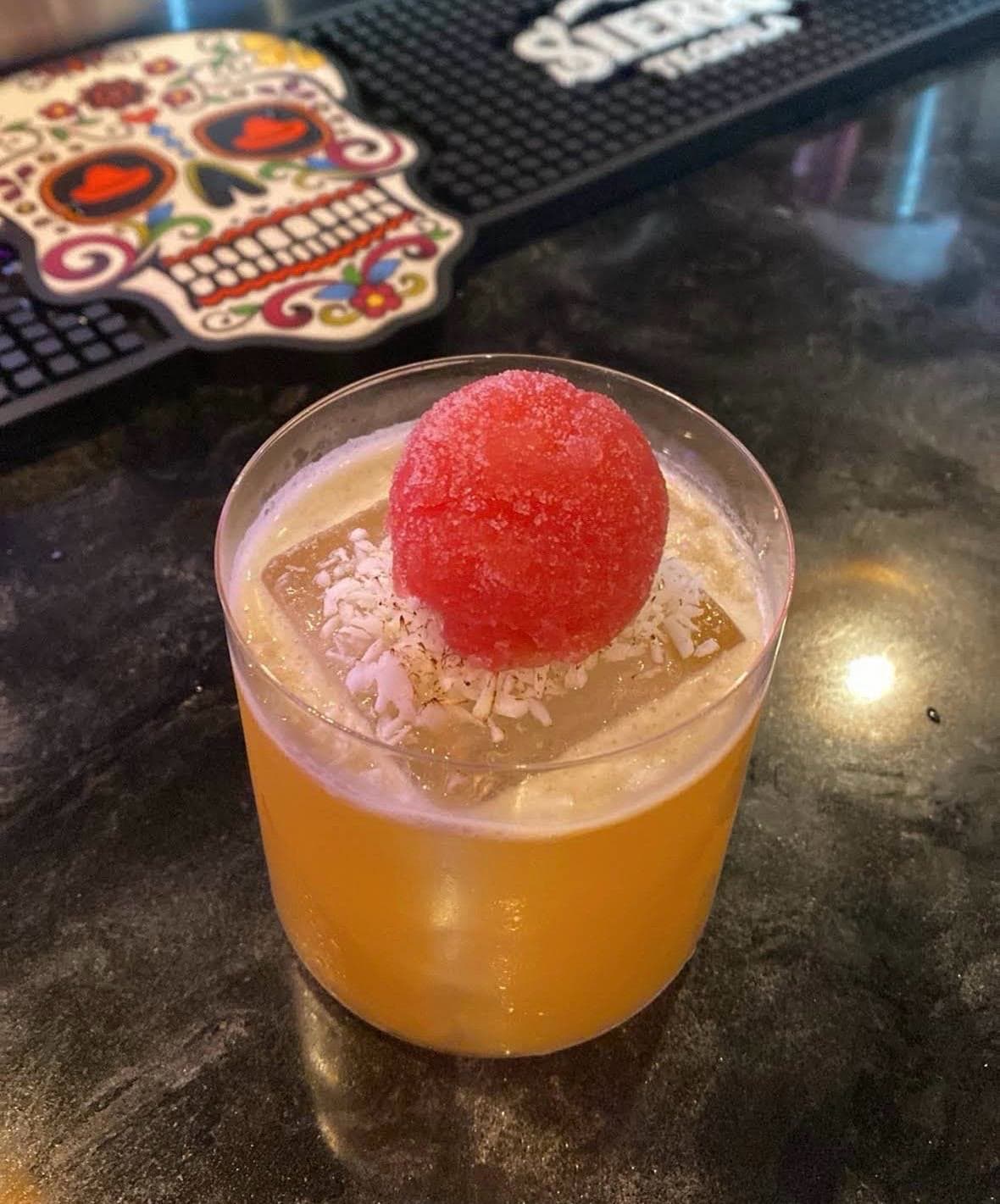 An amber cocktail in a rocks glass topped with a red sugared sphere and shredded coconut, on the dark bar top