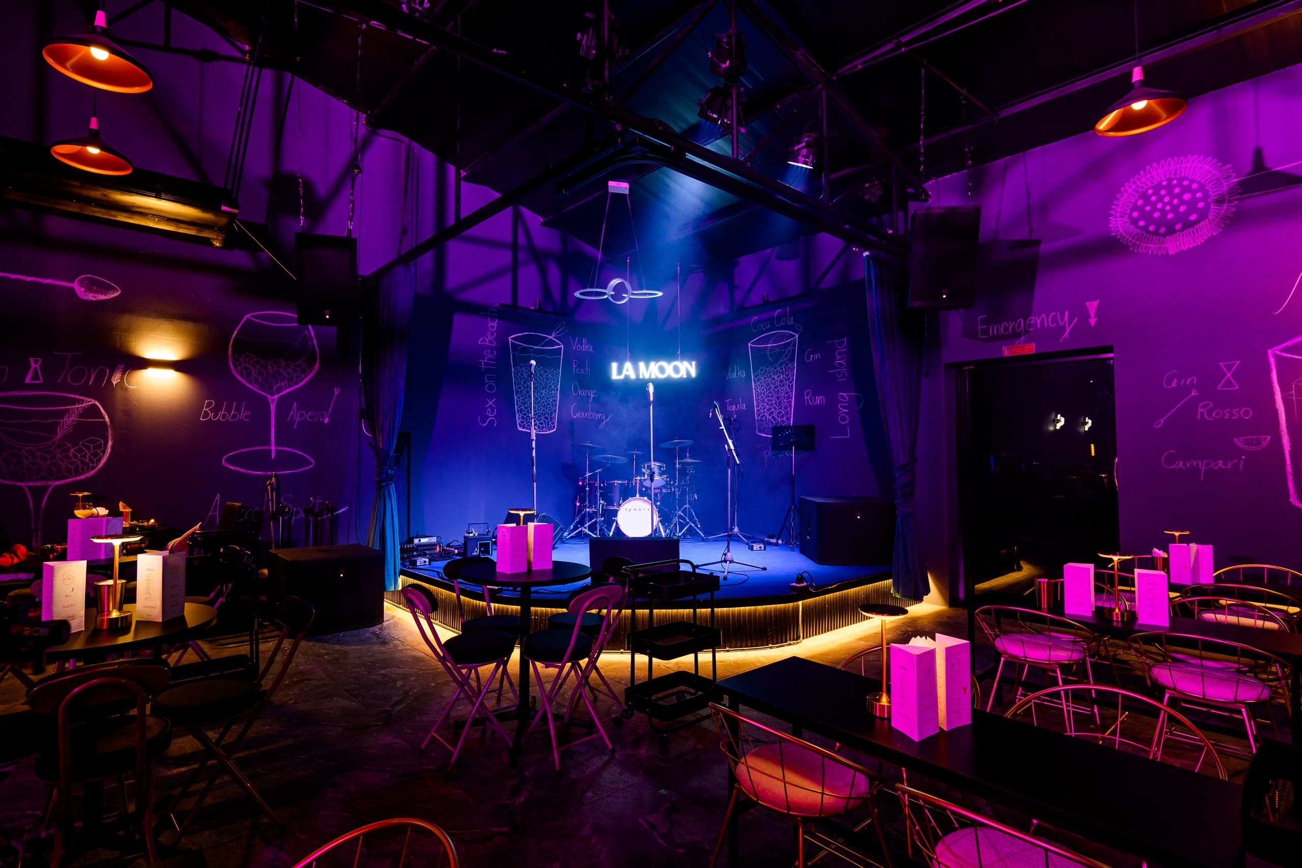 The stage at La Moon under its neon sign, with the hand drawn cocktail wall and tables in front of it
